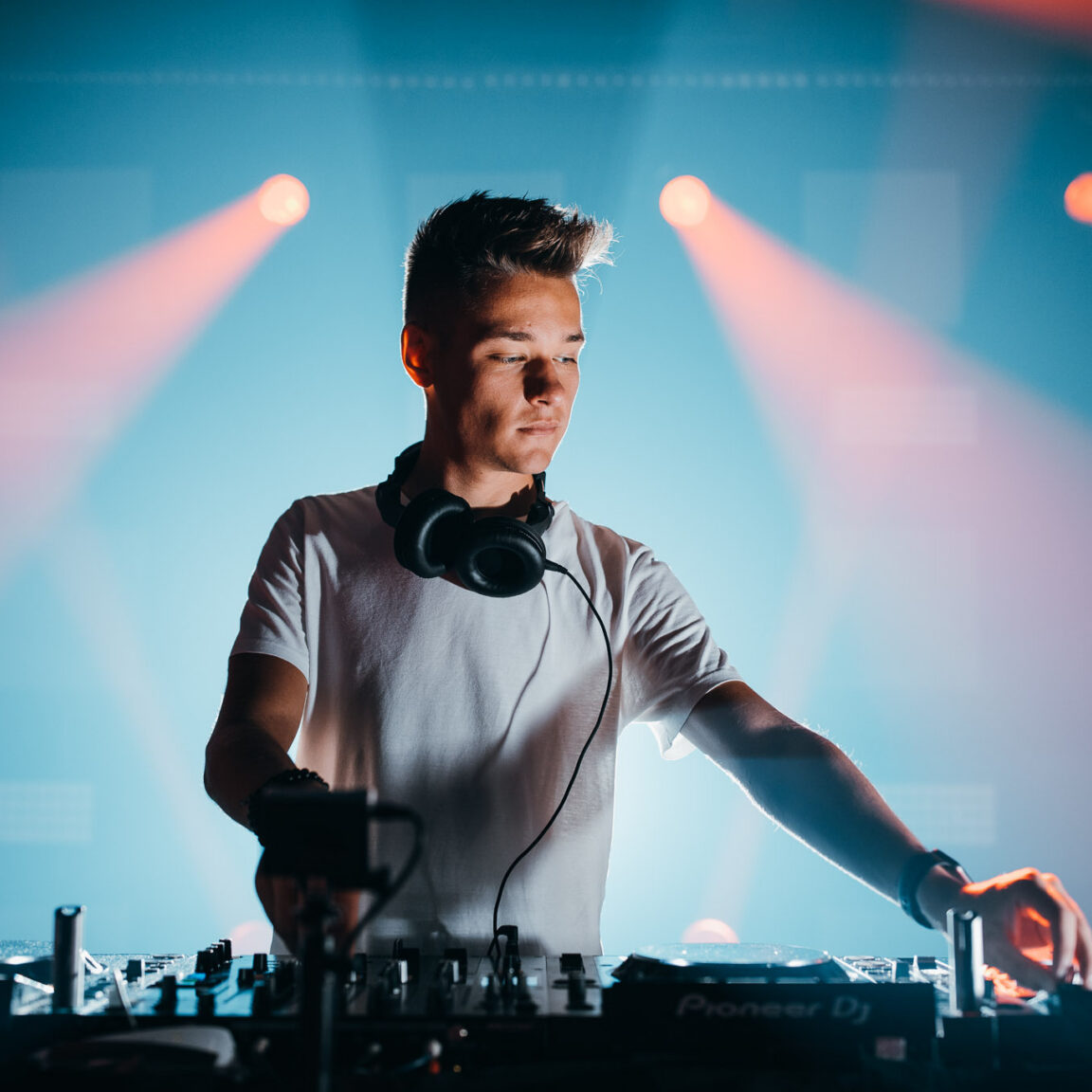 Voltage behind the decks in a white T-shirt, coral and blue stage lights behind him