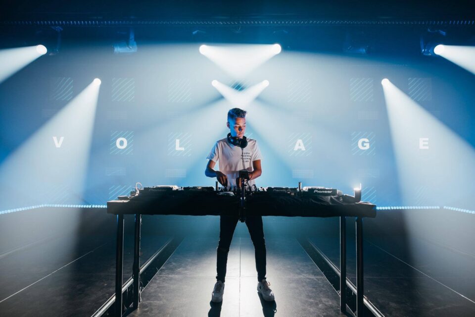 Voltage at the decks under white beams, the letters of VOLTAGE on the screen behind him