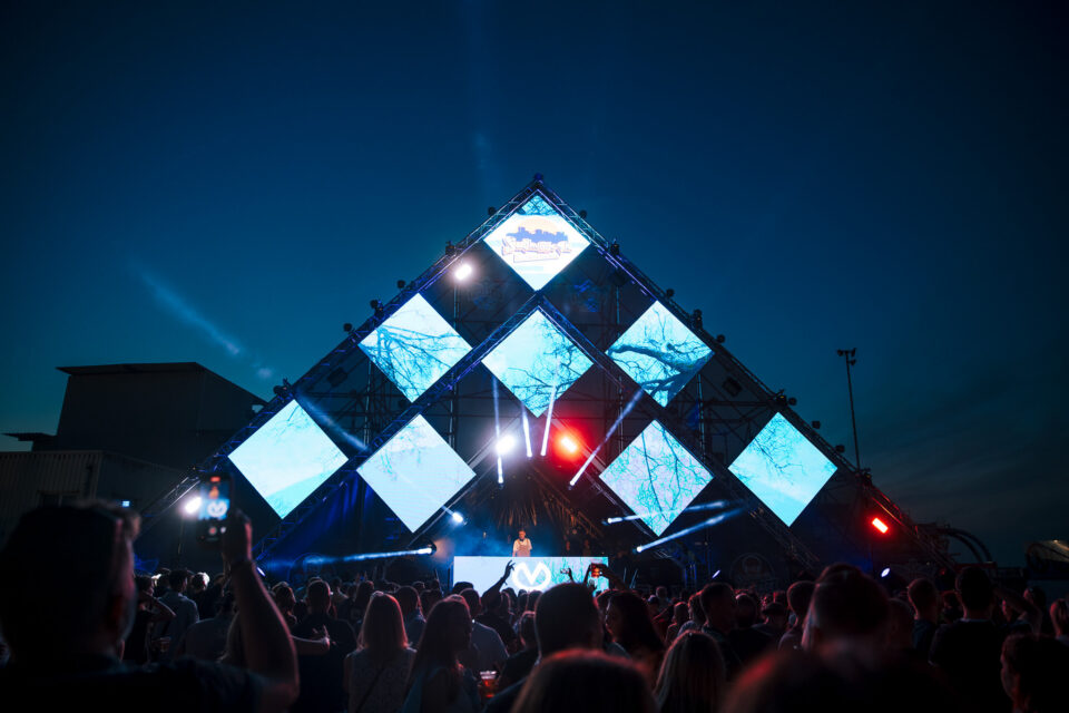 A pyramid stage of diamond-shaped screens at dusk, Voltage at the decks below