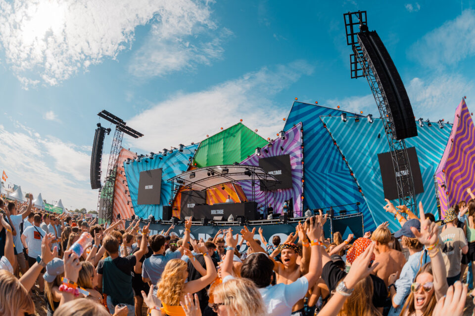 A colourful festival stage with VOLTAGE on the screens and a crowd with hands in the air