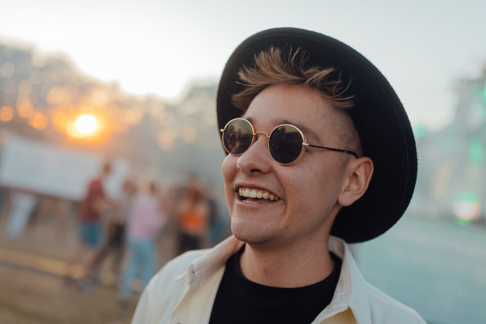 Close-up of Voltage smiling in round sunglasses and a black hat, a festival sunset behind him