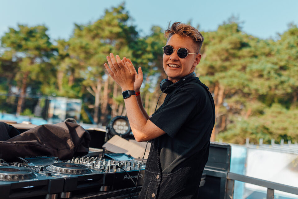Voltage clapping at the decks in round sunglasses, pine trees and blue sky behind