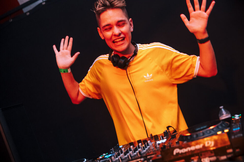 Voltage laughing behind the decks in a yellow shirt, both hands in the air