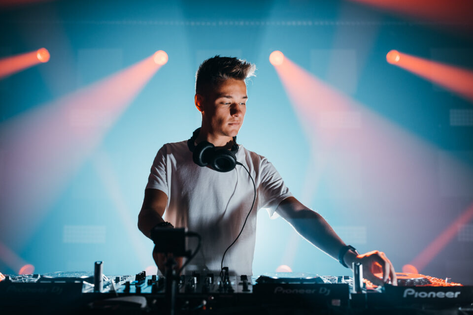 Voltage behind the decks in a white T-shirt, coral and blue stage lights behind him