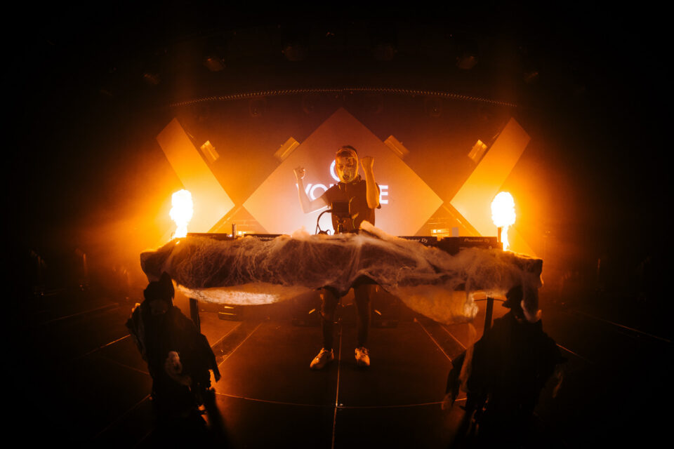 Voltage in a mask with both fists up behind the decks, flames on both sides of the stage