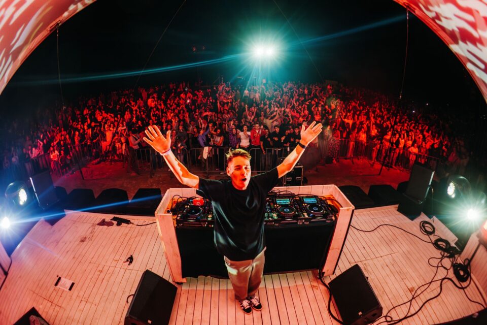 Voltage behind the decks with both arms in the air, facing a packed crowd in red stage light